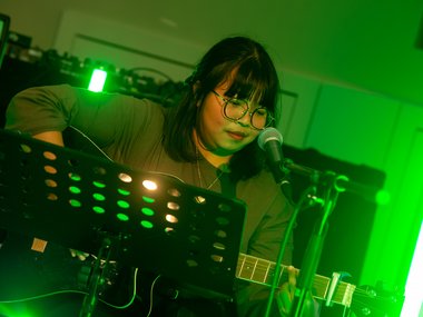 A guitarist performs on stage, surrounded by green lighting. A microphone and music stand with sheet music are in front of the musician.