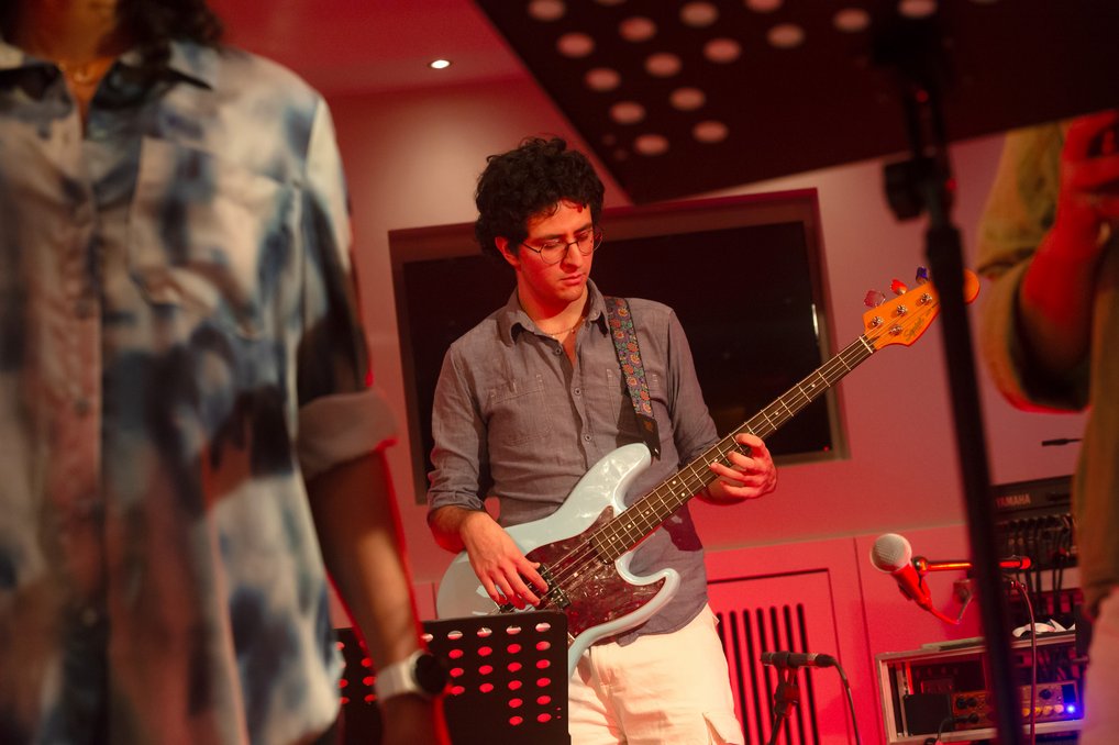 Ein Musiker spielt E-Bass in einem rot beleuchteten Raum, während im Hintergrund ein Mikrofon und weitere Ausrüstung zu sehen sind.