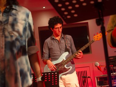 Ein Musiker spielt E-Bass in einem rot beleuchteten Raum, während im Hintergrund ein Mikrofon und weitere Ausrüstung zu sehen sind.