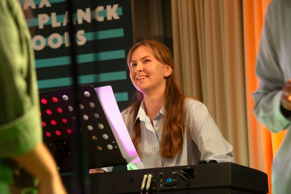 Eine Person in gestreiftem Hemd spielt Keyboard, der beleuchtete Notenständer zeigt Noten. Im Hintergrund ist ein Schild mit dem Text "Max Planck Schools".