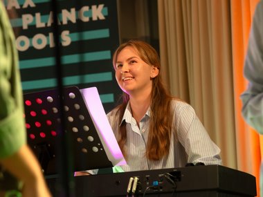 A person plays the keyboard, with a music stand holding sheet music. The background features a banner with the text "Max Planck Schools" and striped design.