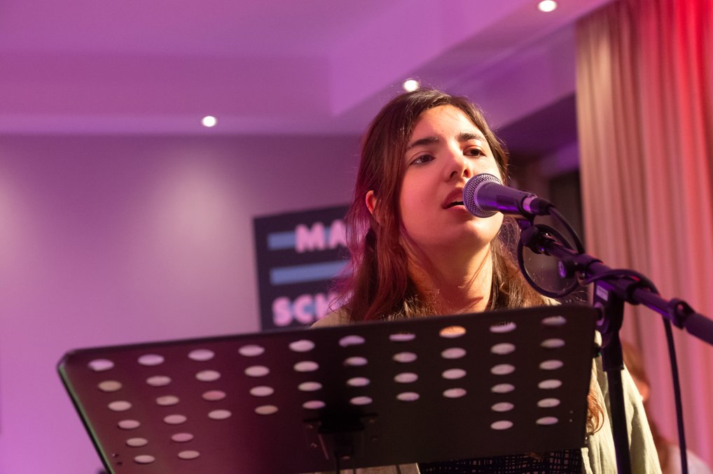 Mikrofon und Notenständer auf einer Bühne, lila und rote Beleuchtung im Hintergrund.