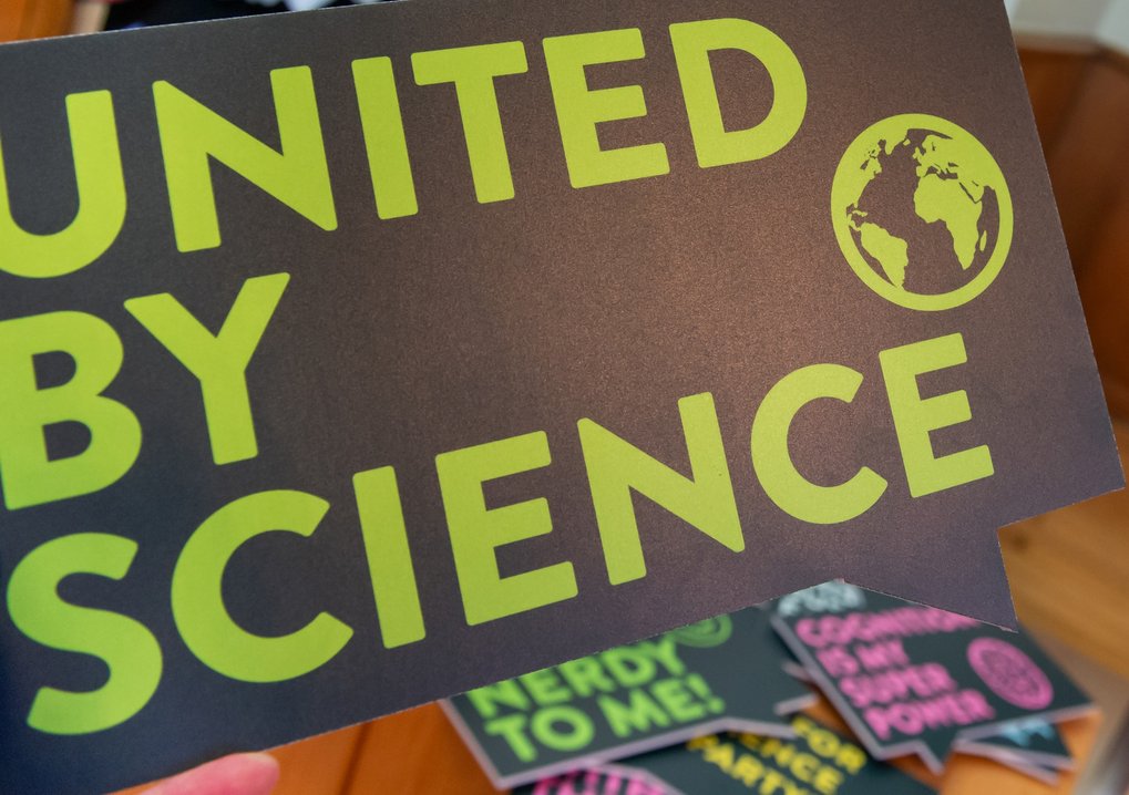Ein Fotostand mit einem Bildschirm, auf dem Fotos angezeigt werden, neben einem Schild mit der Aufschrift 'UNITED BY SCIENCE' in großen grünen Buchstaben. Im Vordergrund sind bunte Requisiten zu sehen, darunter ein Partyhut und ein Schild mit der Aufschrift 'NERDY TO ME!'