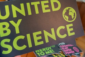Ein Fotostand mit einem Bildschirm, auf dem Fotos angezeigt werden, neben einem Schild mit der Aufschrift 'UNITED BY SCIENCE' in großen grünen Buchstaben. Im Vordergrund sind bunte Requisiten zu sehen, darunter ein Partyhut und ein Schild mit der Aufschrift 'NERDY TO ME!'