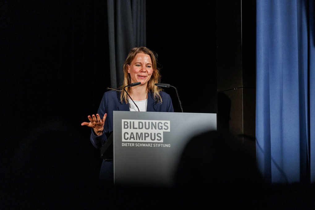 Eine lächelnde Frau steht hinter einem Rednerpult mit der Beschriftung „Bildungs Campus Dieter Schwarz Stiftung“ und spricht vor einem Publikum.
