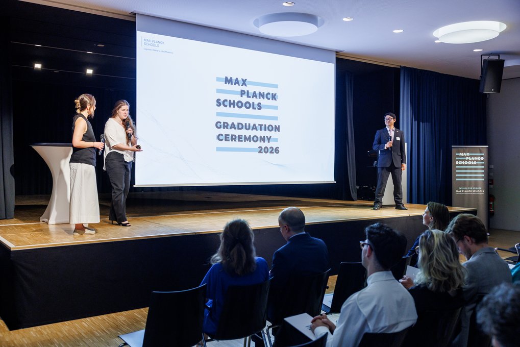 Bei der Abschlussfeier der Max Planck Schools 2026 präsentieren zwei Frauen und ein Mann auf einer Bühne vor einer projizierten Folie mit dem Veranstaltungstitel, während mehrere Personen im Publikum zuhören.