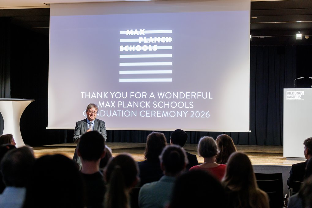 Ein Redner steht vor einer Leinwand mit der Aufschrift „Thank you for a wonderful Max Planck Schools Graduation Ceremony 2026“ vor einem Publikum.