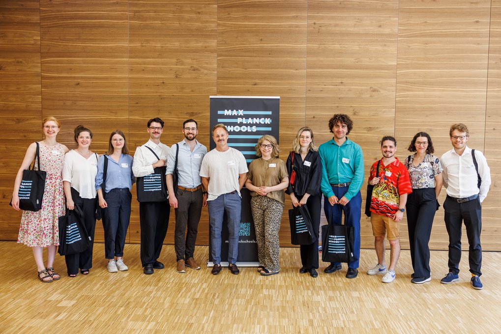Eine Gruppe von elf Personen unterschiedlichen Alters und Geschlechts steht in einem Raum mit Holzverkleidung, einige tragen Taschen mit dem Logo der Max Planck Schools, zentral ein Banner der Organisation.