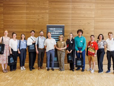 Eine Gruppe von elf Personen unterschiedlichen Alters und Geschlechts steht in einem Raum mit Holzverkleidung, einige tragen Taschen mit dem Logo der Max Planck Schools, zentral ein Banner der Organisation.
