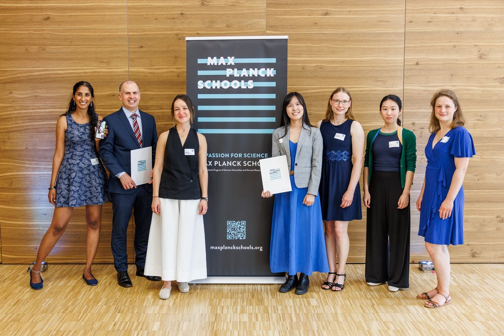 Sieben Personen in formeller und halbformeller Kleidung stehen nebeneinander vor einem Max-Planck-Schools-Banner in einem Raum mit Holzverkleidung und hellen Holzboden.