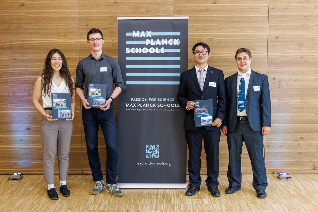 Vier Personen in formeller und semi-formeller Kleidung stehen vor einem Holzpaneel mit einem Banner der Max Planck Schools, auf dem das Logo und die Webseite abgebildet sind, und halten Informationsmaterial.
