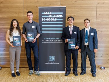 Vier Personen in formeller und semi-formeller Kleidung stehen vor einem Holzpaneel mit einem Banner der Max Planck Schools, auf dem das Logo und die Webseite abgebildet sind, und halten Informationsmaterial.