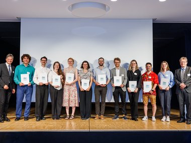 Elf Personen, darunter zwei Männer in Anzügen an den Enden, stehen auf einer Bühne vor einer weißen Leinwand und zeigen Urkunden, vermutlich bei einer Auszeichnung oder Zeremonie.