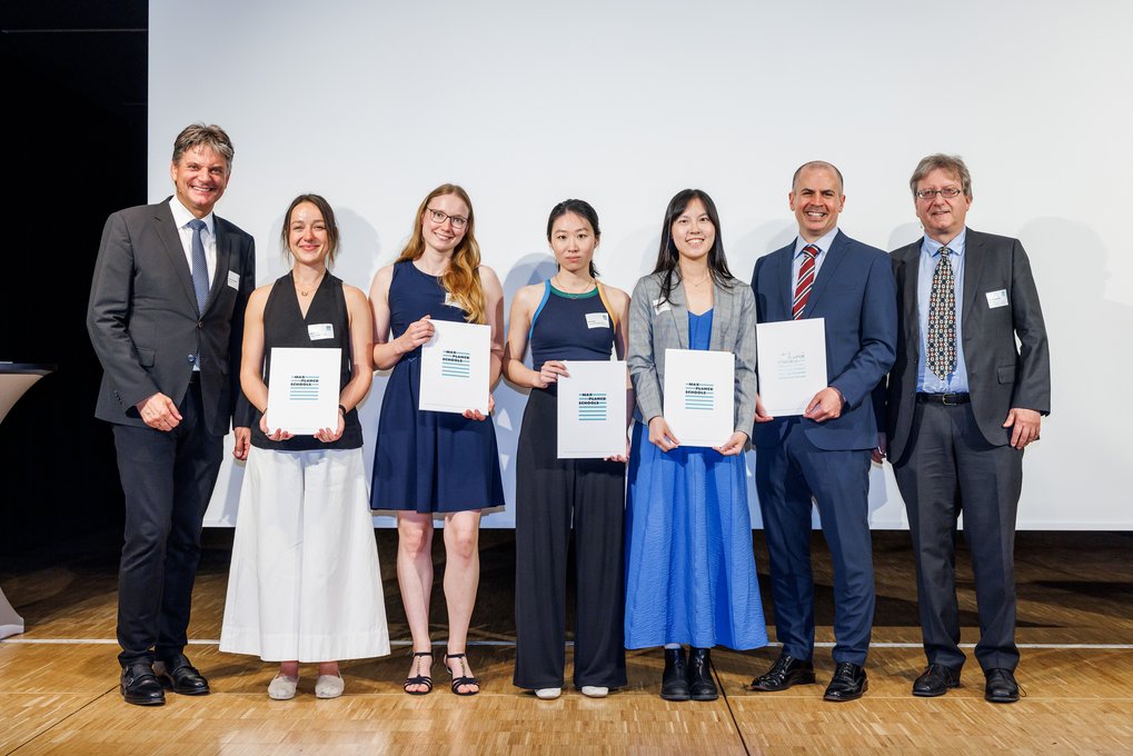 Auf einer Bühne stehen sieben Personen in formeller Kleidung nebeneinander, fünf zeigen Urkunden, die vermutlich Preise oder Zertifikate darstellen, vor einem weißen Hintergrund.