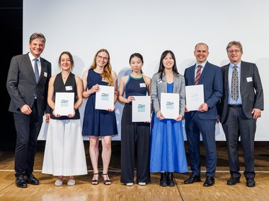 Auf einer Bühne stehen sieben Personen in formeller Kleidung nebeneinander, fünf zeigen Urkunden, die vermutlich Preise oder Zertifikate darstellen, vor einem weißen Hintergrund.
