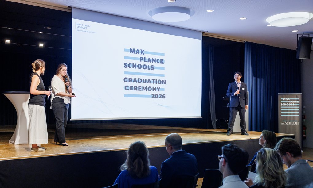 Bei der Max Planck Schools Abschlussfeier 2026 sprechen zwei Frauen und ein Mann auf einer Bühne vor einem sitzenden Publikum, während ein großer Bildschirm die Veranstaltung anzeigt.