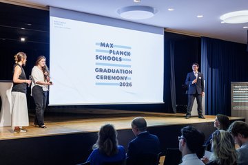 Bei der Max Planck Schools Abschlussfeier 2026 sprechen zwei Frauen und ein Mann auf einer Bühne vor einem sitzenden Publikum, während ein großer Bildschirm die Veranstaltung anzeigt.
