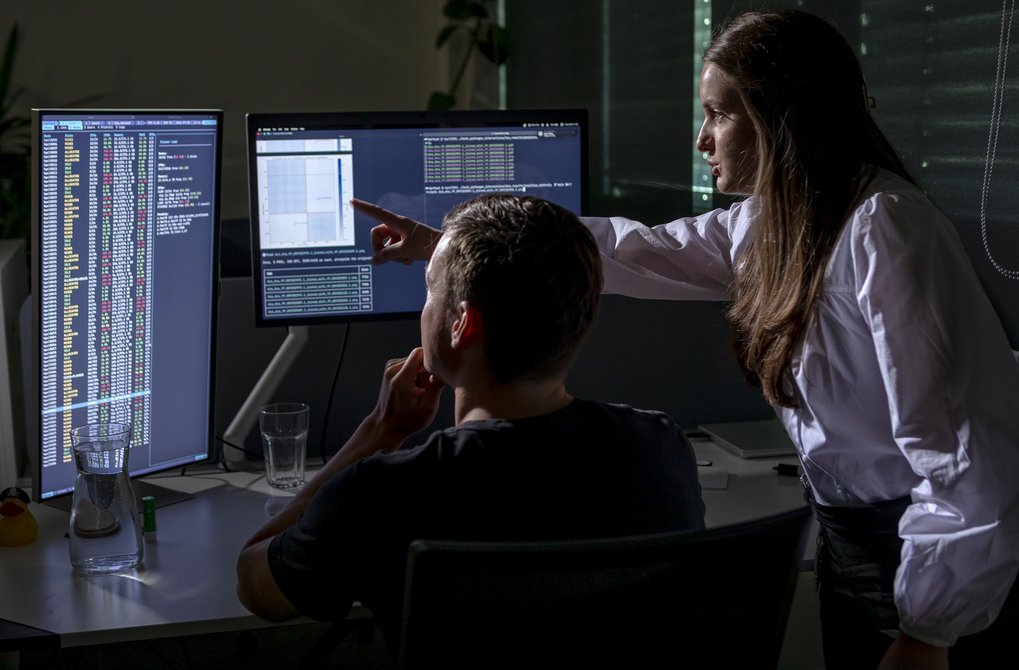 A woman pointing at a computer screen while a man observes data on two monitors in a darkened workspace.