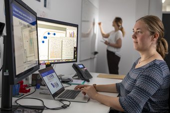 Computergestützte Methoden eröffnen neue Wege in der biomedizinischen Forschung. In einem Büro arbeitet eine Frau an einem Laptop und zwei Monitoren mit Diagrammen und Grafiken, während im Hintergrund eine weitere Frau an einem Whiteboard schreibt.