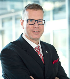 Person wearing a formal business suit, featuring a red striped tie and matching pocket square, positioned in an office setting.