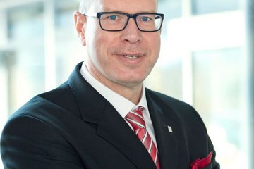 Person wearing a formal business suit, featuring a red striped tie and matching pocket square, positioned in an office setting.